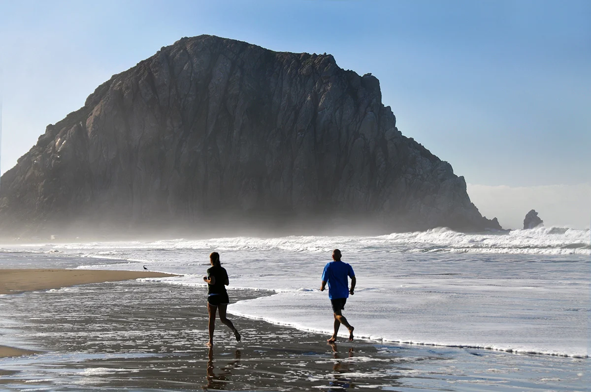Two people jogging on a beach with Morro Rock in the background